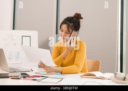 Young adult Asian woman user interface designer discussing mobile application interface wireframe on a phone call brainstorming user experience planni Stock Photo