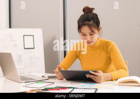 Asian adult woman UX UI designer working on tablet device creating innovative interface designs for mobile app or website development. Focus on user e Stock Photo