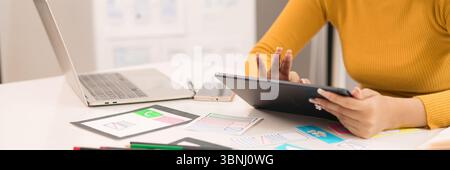 An Asian adult woman's hands are focused on designing a user interface using a tablet near a laptop and mobile phone. She is creating a design for a w Stock Photo