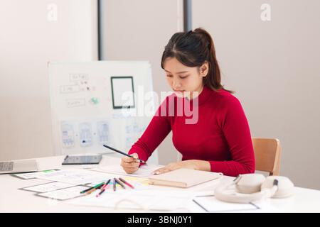 Young Asian woman user interface designer is working on mobile application design at her desk with sketches. She designs website project and brainstor Stock Photo