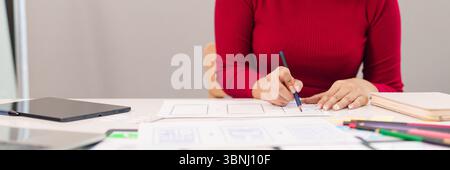 Adult Asian woman uses pencil to sketch user interface designs on paper. UI UX designer at work developing mobile app layouts for new startup project. Stock Photo