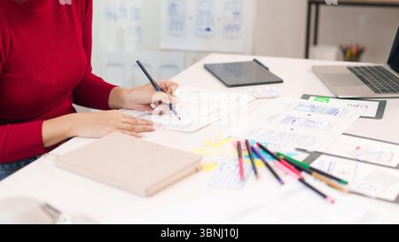 An Asian adult women creating web design on paper. The user interface designer is at work, developing mobile app with hand sketching wireframe. Stock Photo