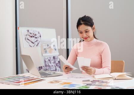 An adult Asian woman UI UX designer reviews design documents and brainstorms ideas for a user interface design project using her laptop and color pale Stock Photo