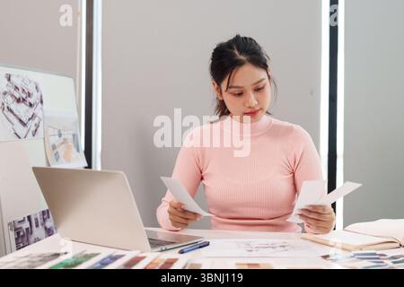 Young Asian woman is a UI UX designer working at her desk with laptop and color palette. She's reviewing design documents. Creative inspiration at wor Stock Photo