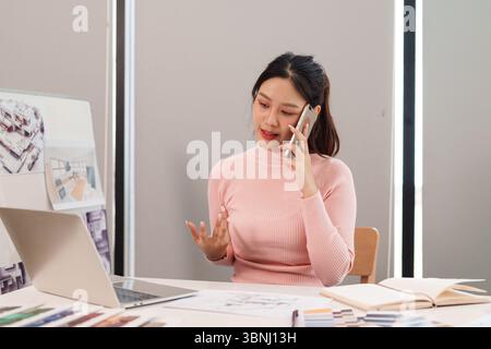 An adult Asian woman UI UX designer is speaking on her smartphone during her work in the office, showcasing her design process, user interface and exp Stock Photo