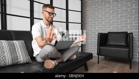 Young bearded man with laptop meditating on couch in office Stock Photo