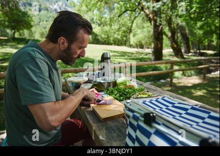 A man chops fresh vegetables on a wooden table in a scenic park. Surroundings include trees, a picnic table, and culinary items, emphasizing outdoor c Stock Photo