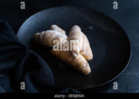On a dark plate, homemade golden croissants with a light dusting of flour are arranged. The rustic presentation enhances their fresh, delicious appeal Stock Photo