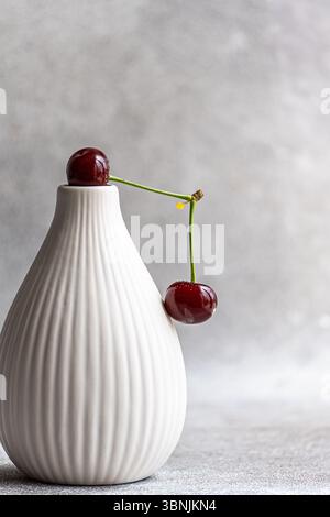 A ripe cherry delicately rests on an elegant white ceramic vase, with a soft concrete background adding texture to this minimalistic and stylish compo Stock Photo
