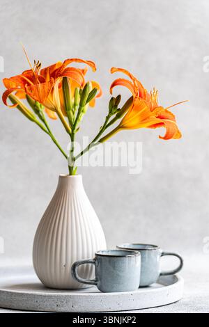 Vibrant orange tiger lilies elegantly arranged in a white vase, placed on a minimalist circular tray alongside two ceramic cups against a soft gray ba Stock Photo