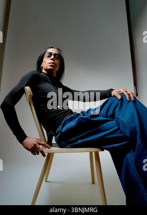 A portrait of a stylish Latin man sitting on a wooden chair, wearing a sleek black long-sleeve shirt and blue jeans. His confident pose exudes casual Stock Photo