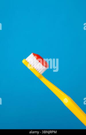 A vibrant yellow plastic toothbrush with soft bristles holds a dab of shiny red toothpaste. The toothbrush stands out vividly against a solid blue bac Stock Photo