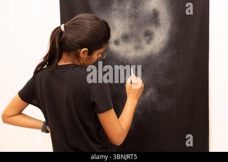 A young Indian woman artist, focused intently, uses a white pencil to create art on a dark canvas. Her concentrated expression reflects her passion an Stock Photo