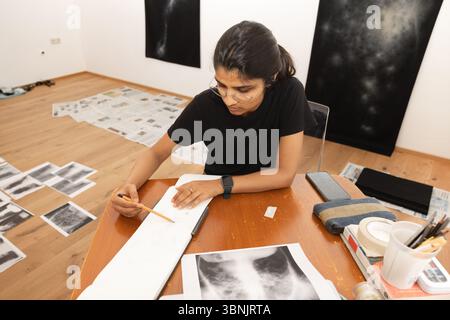 A young Indian woman artist engaged in sketching at her home studio, surrounded by her artworks and materials. She is focused, wearing glasses and a s Stock Photo