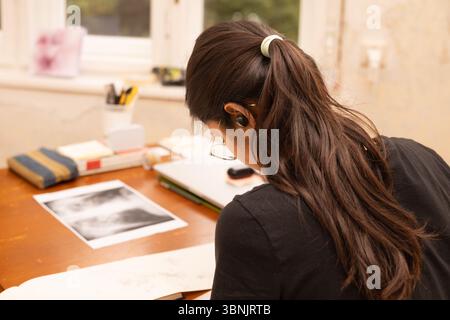 A young Indian woman artist is focused on her creative work in a cozy studio setting. Surrounded by art supplies, she concentrates intently on her ske Stock Photo