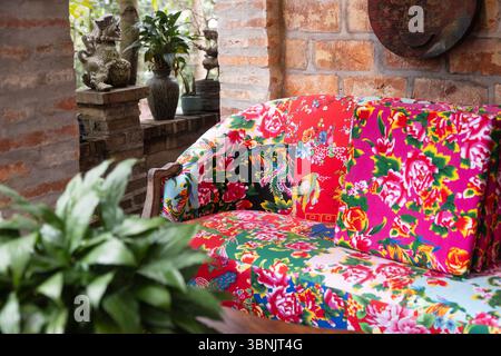 Colorful floral patterned sofa with vibrant cushions in a traditional Vietnamese patio setting, surrounded by rustic brick walls and lush potted plant Stock Photo