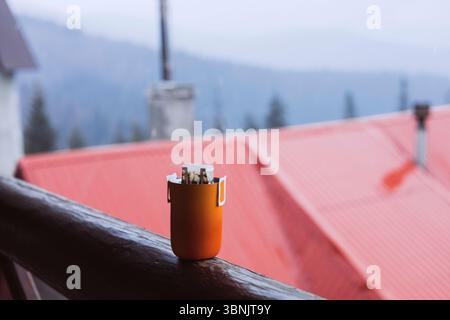 Orange coffee cup with paper filter sits on a wooden railing, overlooking misty mountains and red roofs, evoking a calm and serene atmosphere in a mou Stock Photo