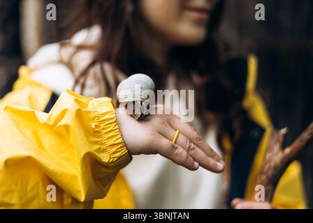 A young woman in a vibrant yellow raincoat gently holds a wooden stick while a snail rests on her hand, surrounded by the lush greenery of a mountaino Stock Photo