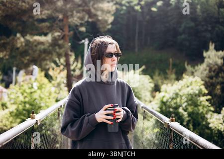 A young woman in a dark hoodie stands on a bridge, holding a thermos cup. She is wearing sunglasses and surrounded by lush greenery, enjoying a serene Stock Photo