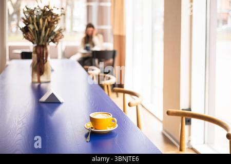 A vibrant yellow coffee cup with saucer and spoon sits prominently on a sleek blue table in a modern cafe, with blurred patrons in the background enjo Stock Photo