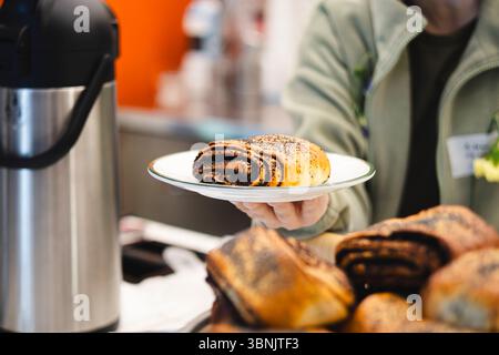 Cropped unrecognizable coffee shop worker offers a poppy seed bun on a plate. The fresh pastry contrasts with the background, creating a welcoming atm Stock Photo