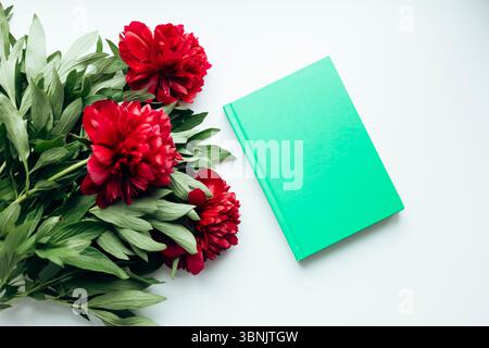 A lush bouquet of red peonies arranged on a table next to a closed green book. This elegant indoor setting captures the beauty and freshness of natura Stock Photo