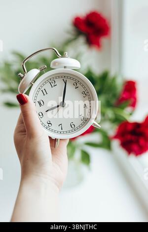 A close-up of a hand holding a classic white alarm clock, with vibrant red peonies in the blurred background, evoking a sense of holiday relaxation an Stock Photo
