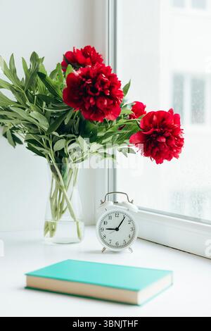 A serene scene with a bouquet of vibrant red peonies in a glass vase beside a classic white alarm clock on a windowsill. A teal book adds a subtle pop Stock Photo