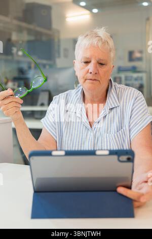 An elderly woman sits at home, using a tablet with focus and contemplation. She seems engaged and thoughtful, enhancing her digital literacy and stayi Stock Photo