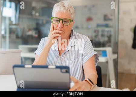 Thoughtful elderly woman wearing eyewear, sitting at home and using a tablet. She appears to be engaged in deep thought, possibly reading or researchi Stock Photo