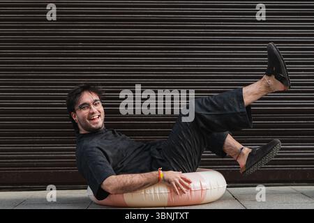 Smiling man in glasses and black attire sits on a pink inflatable in front of a dark, modern shutter. His casual summer style and joyful demeanor depi Stock Photo