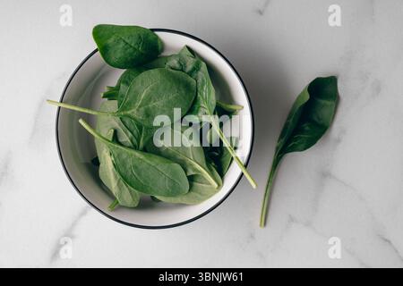 Top view of fresh spinach leaves in a white bowl placed on a marble surface. The vibrant green colors of the spinach highlight their freshness, making Stock Photo