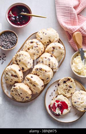Top view of freshly baked scones with chocolate chips, served with clotted cream and cherry jam on a rustic table setting, surrounded by dessert ingre Stock Photo