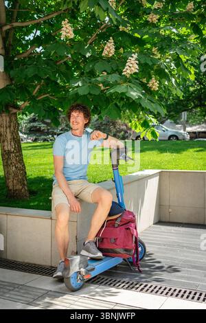 A man sits on a concrete ledge in a park, leaning on his electric scooter. He enjoys the sunny day under the leafy shade of a tree, looking relaxed an Stock Photo