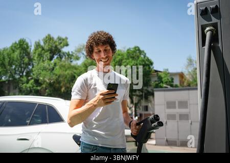 A man happily charges his electric vehicle at a public charging station on a sunny day, checking his smartphone. Green trees and a bright sky frame th Stock Photo