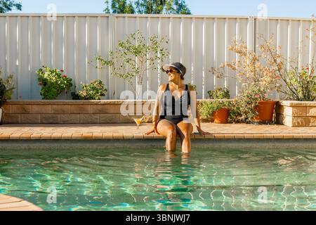 Woman in swimsuit, sun visor hat and sunglasses relaxing sitting at the edge of a sunny pool, enjoying the ambiance. She is smiling and looking conten Stock Photo