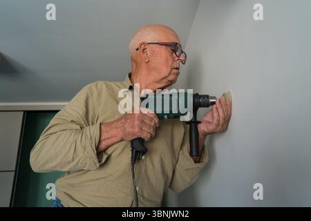An elderly man wearing glasses is focused on a DIY project at home, using a power drill to work on a wall, showcasing skill and dedication in home imp Stock Photo