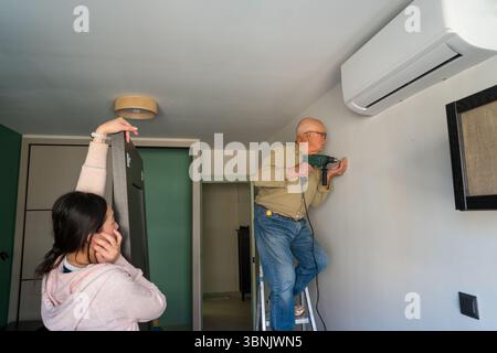 An elderly man using a power drill while standing on a ladder, assisted by a young woman holding a door. They both are engaged in a DIY home improveme Stock Photo
