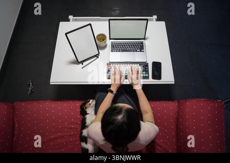 An Asian woman works remotely from home on her laptop, multitasking efficiently while taking care of her adorable puppy sitting beside her on the couc Stock Photo