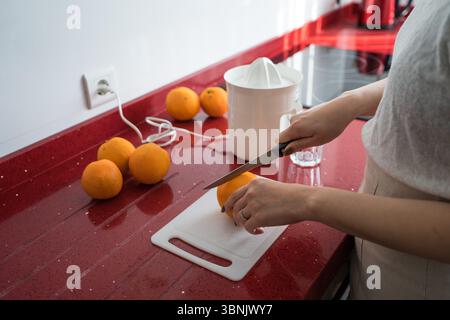 Cropped image of a woman’s hands preparing fresh orange juice in a sleek modern kitchen using a juicer. Oranges and a cutting board are placed on a st Stock Photo