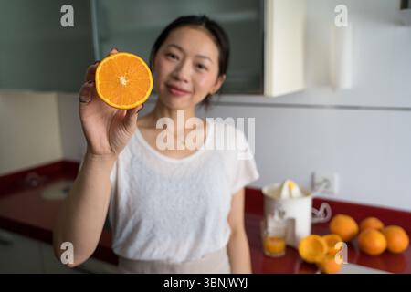 Young Asian woman smiles at the camera while preparing fresh orange juice in a modern kitchen, highlighting a healthy lifestyle with fresh ingredients Stock Photo