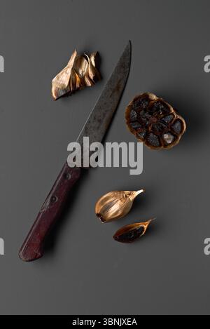 A vintage knife with a wooden handle lies alongside peeled black garlic cloves on a dark surface. The rustic textures of the knife contrast with the g Stock Photo
