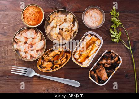 Top view of various canned seafood, including mussels, clams, and sardines, neatly arranged on a rustic wooden table. A fork and parsley garnish compl Stock Photo