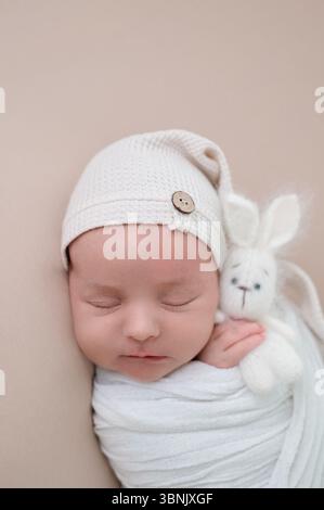 A serene newborn baby peacefully sleeps wrapped in a cozy white blanket, snuggling a small plush bunny. The baby wears a cute knitted hat, enhancing t Stock Photo