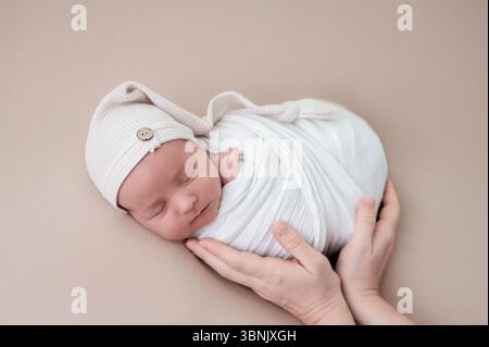 A serene image of a sleeping newborn baby wrapped snugly in a soft white blanket, wearing a cute knitted hat, surrounded by gentle, protective hands Stock Photo