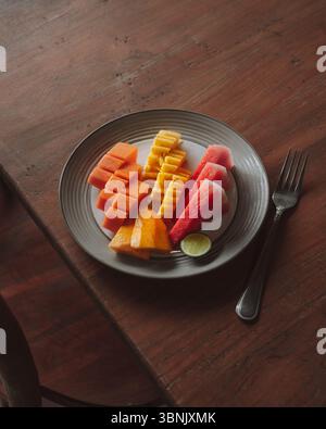 A beautifully arranged plate of tropical fruits including papaya, pineapple, watermelon, and a slice of lime, set on a wooden table in a cozy hotel di Stock Photo