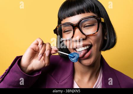 Hispanic young woman with short hair and large glasses enjoying a blue heart-shaped lollipop. She's wearing a purple blazer and smiling against a vibr Stock Photo