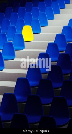 A vivid yellow stadium seat sits alone among a sea of blue chairs, creating a striking visual contrast in an organized setting. Stock Photo