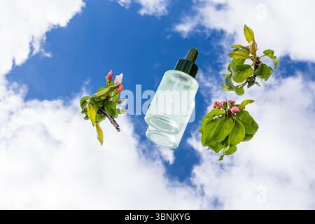 A serum bottle is reflected on a mirror, set against a vibrant blue sky with fluffy white clouds. A branch with fresh leaves and small flowers complem Stock Photo