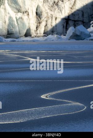 A stunning winter scene in Iceland, showcasing intricate patterns of frozen ice on a lake, with rugged cliffs and snow in the background. Stock Photo